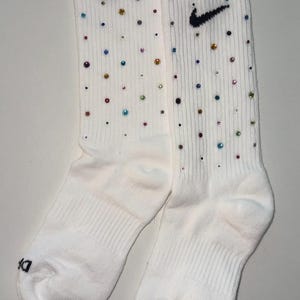 May include: A pair of white athletic socks adorned with colorful rhinestones and a black Nike swoosh logo. The socks have a ribbed design and the letters "DR" are printed on the cuff.