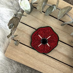 May include: A red beaded poppy flower with a black centre and white stitching. The poppy is on a wooden surface.