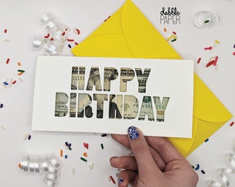 May include: A white card with the words "Happy Birthday" cut out of the card. The cut-out letters are filled with US dollar bills.