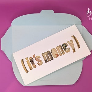 May include: A white card with the words "it's money" cut out of the card, revealing dollar bills behind the cutouts. The card is sitting inside a light blue envelope.