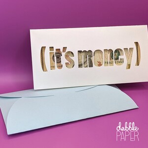 May include: A white card with the words "(it's money)" cut out, revealing a dollar bill. The card is sitting on top of a light blue envelope. The text "dabble PAPER" is printed in white on the bottom right corner of the envelope.