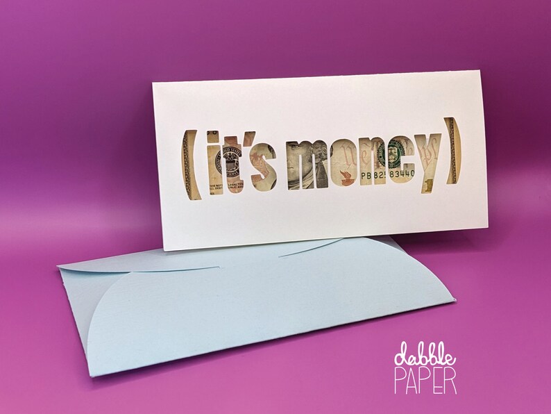 May include: A white card with the words "(it's money)" cut out, revealing a dollar bill. The card is sitting on top of a light blue envelope. The text "dabble PAPER" is printed in white on the bottom right corner of the envelope.
