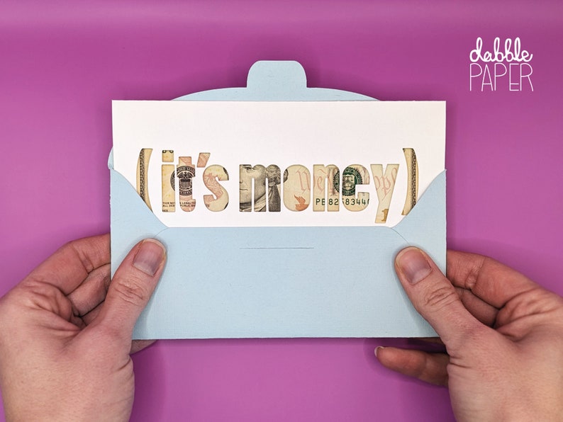 May include: A light blue envelope with a white card inside. The card has the words "it's money" cut out of it, and the cut-out letters are filled with US dollar bills.