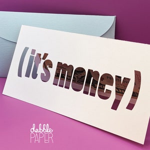 May include: White card with the words "(it's money)" cut out, revealing a dollar bill underneath. The card is folded in half and has the words "dabble PAPER" printed on the bottom.