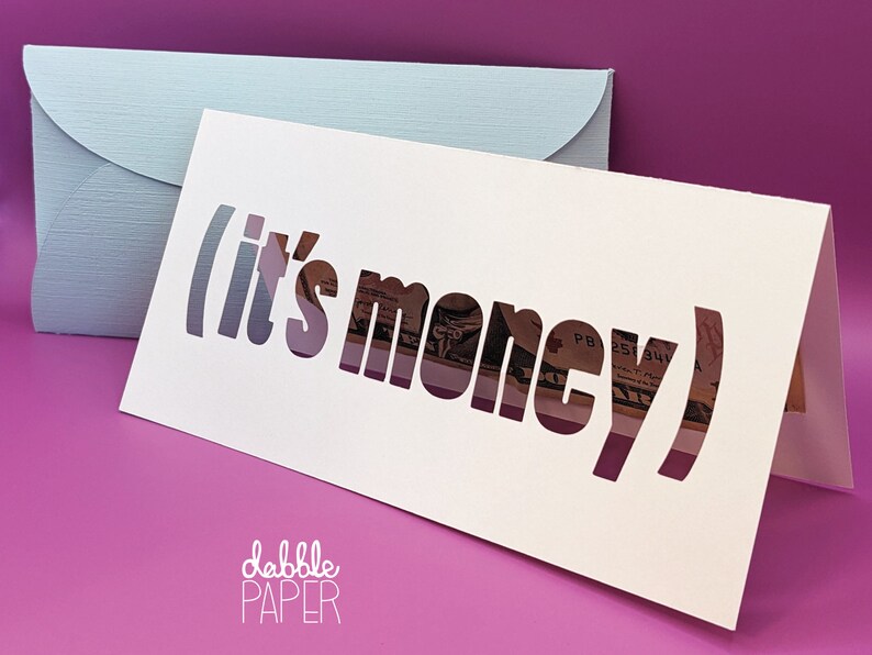 May include: White card with the words "(it's money)" cut out, revealing a dollar bill underneath. The card is folded in half and has the words "dabble PAPER" printed on the bottom.