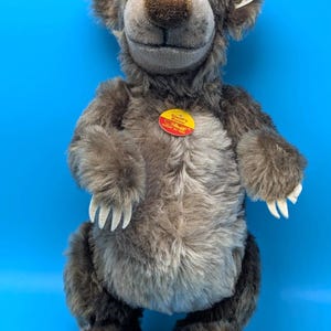May include: A brown plush bear toy with shaggy fur, a lighter tan chest, and white felt claws. The bear features a smiling expression, large eyes, and a red circular chest tag that reads Steiff's Baloo, with a white tag attached to the ear.