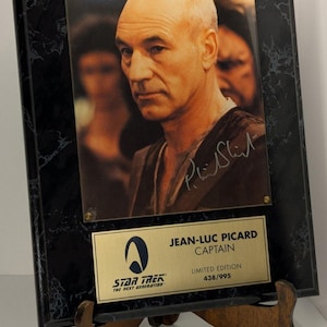 May include: A framed Star Trek: The Next Generation collectible featuring Jean-Luc Picard, Captain of the USS Enterprise. The image is mounted on a black marble-patterned frame with a gold plaque that reads "Limited Edition 438/995".