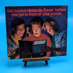 May include: A vintage Nintendo Power magazine advertisement featuring three individuals and a dog looking at the publication. The text reads: Get behind Nintendo Power before you get in front of your screen. The magazine cover shows Mario.