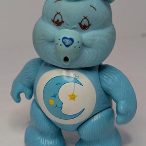 May include: A light blue Care Bear figurine. The bear has a crescent moon and star design on its belly. The bear has a blue nose with a heart, and blue hair. The bear is a collectible toy.