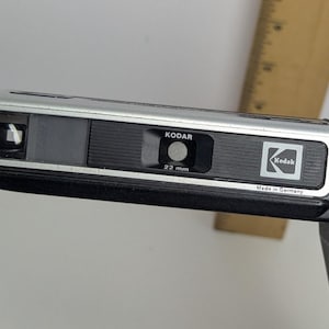 May include: A vintage Kodak Kodar 22 mm camera with a silver and black body. The camera has a black strap and the Kodak logo is visible. The camera is marked "Made in Germany".