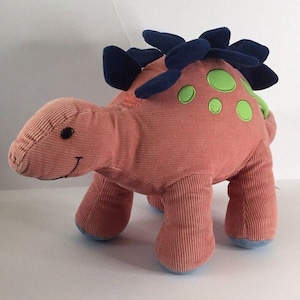 May include: A plush dinosaur toy in a coral corduroy fabric. It features dark blue dorsal plates, green spots, and blue accents on the feet. The dinosaur has a smiling face and is designed for children.