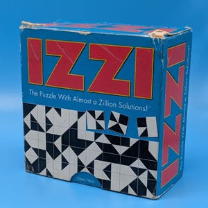 May include: A blue cardboard game box for the Izzi puzzle. The front features the red text IZZI, the subtitle The Puzzle With Almost a Zillion Solutions!, and a grid of black and white geometric tile patterns. The side also displays the red IZZI text.