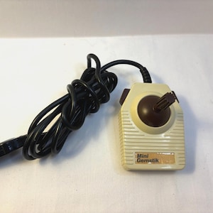 May include: A vintage Mini Gemstik joystick controller. The beige plastic controller has a brown joystick and a black cable. The text "Mini Gemstik" is visible on the front. The cable is coiled.