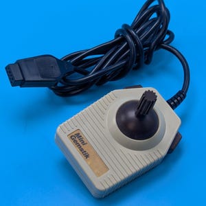 May include: A beige video game joystick controller with a black stick and a black button on the side. The device features a label that reads Mini Gemstik and includes a black coiled cable with a rectangular connector plug at the end.