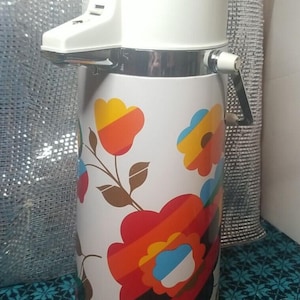 May include: A vintage white thermos with a floral design in orange, red, yellow, and blue. The thermos has a white top with a spout and a chrome handle. The floral pattern is on a white background.