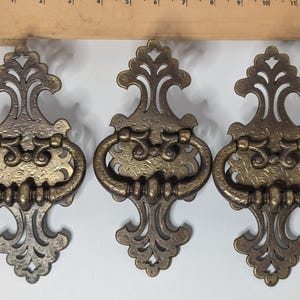 May include: Three antique brass drawer pulls with ornate, scrolling designs. Each pull features a decorative handle and a detailed backplate. The pulls are in varying shades of gold and bronze, with a vintage aesthetic.