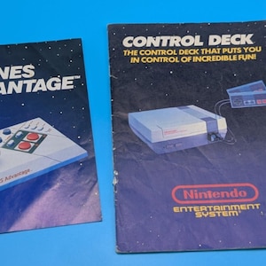 May include: Two vintage Nintendo instruction manuals with dark blue space-themed covers. The left manual features the NES Advantage controller, while the right manual displays the Nintendo Entertainment System console with the text Control Deck.