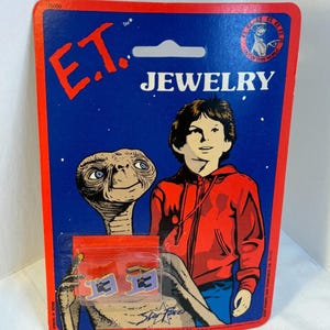May include: Vintage E.T. the Extra-Terrestrial jewelry set on a blue and red card. The card features the movie's logo and characters, with the word "JEWELRY" in white. The jewelry is in a small plastic bag.
