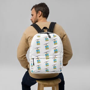 May include: White backpack with a repeating pattern of colorful ski lift gondolas. The backpack has black shoulder straps and a black zipper. The ski lift gondolas are blue, yellow, and green.