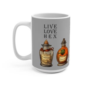 May include: A white ceramic mug with a gray background featuring the text "LIVE LOVE HEX" and illustrations of potion bottles. The bottles are in shades of brown, orange, and gold, with labels and corks.