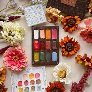 Könnte beinhalten: Eine rechteckige Aquarellpalette aus Holz mit zwölf rechteckigen Näpfchen aus fester Farbe in verschiedenen Tönen, begleitet von einem dunklen Holzdeckel mit Lederlogo und zwei gedruckten Pigment-Referenzkarten, umgeben von bunten Blumen.