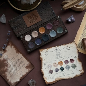 May include: A dark wooden watercolor paint palette with ten circular pans of pigment in various colors, featuring a leather label with a rabbit logo. Includes a textured paper swatch card labeled Foxy Wolf with color names and decorative ink borders.