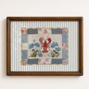 May include: A rectangular wooden frame containing a fabric collage with a central embroidered red lobster flanked by blue flowers. The piece features a patchwork border of blue gingham and pink floral fabric, set against a blue and white striped background.