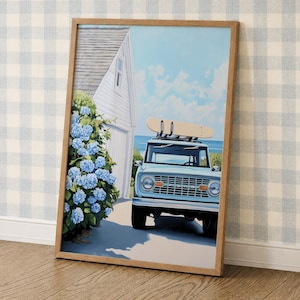 May include: A framed wall art print depicting a light blue vintage SUV with a surfboard on its roof rack, parked next to a white house with a large bush of blue hydrangea flowers. The scene features a clear blue sky and a coastal background.