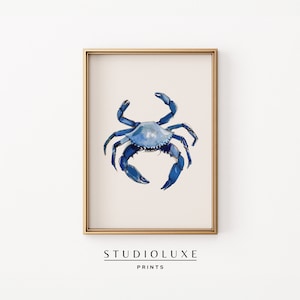 May include: A framed print featuring a watercolor illustration of a blue crab. The crab is centered on a cream-colored background, with a gold-colored frame. The text "STUDIOLUXE PRINTS" is at the bottom.