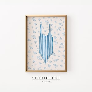 May include: A framed print of a blue and white striped swimsuit on a light pink background with a white floral pattern. The print is framed in a gold frame. The text "STUDIO LUXE PRINTS" is below the frame.