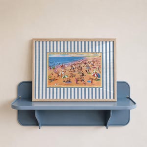 Könnte beinhalten: Ein gerahmter Vintage-Postkarten-Druck einer belebten Strandszene mit Menschen, Sonnenschirmen und dem Meer. Der Rahmen hat einen blau-weiß gestreiften Rand und wird auf einem blauen Regal ausgestellt. Der Postkartentext lautet "Surf Bathing at Old Silver Beach, West Falmouth, Mass."