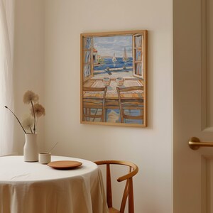May include: A framed painting of a seascape with two sailboats in the distance. The painting depicts a table with two chairs and two cups of coffee. The painting is in a light wood frame.