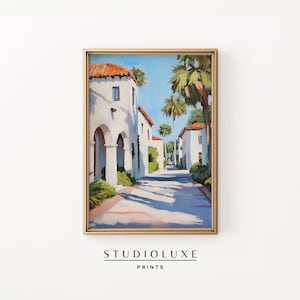 Puede incluir: Una impresión artística enmarcada de una escena callejera soleada. Edificios blancos con tejados de tejas rojas bordean la carretera, con palmeras y un cielo azul. La impresión está en un marco dorado, con el texto "STUDIOLUXE PRINTS" en la parte inferior.