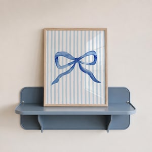 May include: A framed watercolor print featuring a blue bow on a striped background of white and light blue. The artwork is displayed on a blue shelf, creating a cohesive and decorative display. The frame is made of light wood.