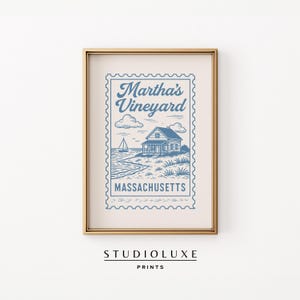 Op de afbeelding: Een ingelijste print met een blauw-witte illustratie van Martha's Vineyard, Massachusetts, ontworpen als een postzegel. De afbeelding toont een kustlandschap met een huis, zeilboot en de woorden "Martha's Vineyard" en "Massachusetts".