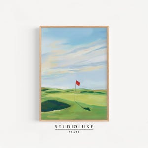 Puede incluir: Impresión artística enmarcada de una escena de campo de golf. La pintura representa un campo de golf verde con una bandera roja, bajo un cielo azul con nubes. La impresión está etiquetada "STUDIOLUXE PRINTS" en la parte inferior.