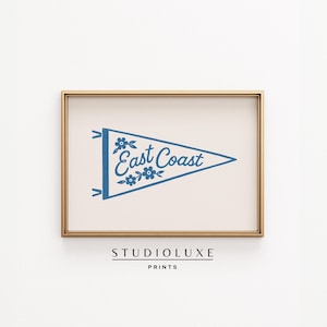 Puede incluir: Una impresión enmarcada con un diseño de banderín azul, que muestra las palabras "East Coast" y detalles florales. La impresión está sobre un fondo crema, encerrada en un marco dorado. El texto "STUDIOLUXE PRINTS" está en la parte inferior.