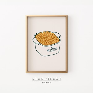Puede incluir: Una impresión enmarcada que presenta una fuente para horno llena de macarrones con queso. La fuente es blanca con un contorno verde azulado y detalles florales. Los macarrones son de color marrón dorado. La impresión está sobre un fondo crema, con el texto "STUDIOLUXE PRINTS" en la parte inferior.