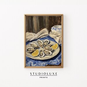 May include: A framed print of a still life painting featuring oysters on a plate with lemon wedges. The plate sits on a blue tablecloth, with a white cloth and a small bowl in the background. The frame is gold, and the print has the text "STUDIOLUXE PRINTS" at the bottom.