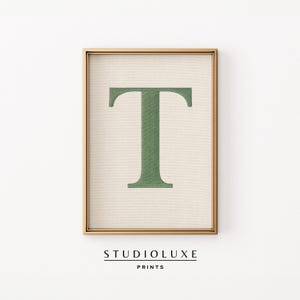 Puede incluir: Impresión enmarcada con la letra "T" en un tono verde apagado. La letra está centrada sobre un fondo beige, dentro de un marco dorado. El texto "STUDIOLUXE PRINTS" se muestra en la parte inferior.