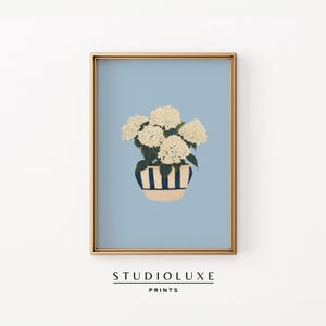 May include: A framed print featuring a light blue background with a gold frame. The artwork depicts a white hydrangea plant in a blue and white striped pot. The text "STUDIOLUXE PRINTS" is at the bottom.