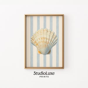May include: Framed art print featuring a detailed seashell illustration. The shell is cream and gold, set against a backdrop of vertical light blue and cream stripes. The frame is gold, and the print includes the text "StudioLuxe PRINTS".