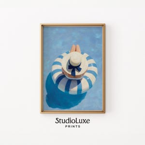 May include: A framed print featuring a person relaxing in a pool on a blue and white striped inflatable ring. The person wears a white hat with a blue bow. The artwork is signed "StudioLuxe PRINTS".