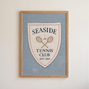 May include: A framed wall art piece featuring a cream-colored fabric shield with red stitched edges. The text SEASIDE TENNIS CLUB EST. 1964 is embroidered in navy blue, centered above and below two crossed tennis rackets and a small sailboat.