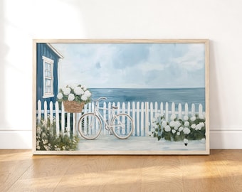 May include: A framed painting depicts a coastal scene. A white bicycle with a basket of white flowers sits near a white picket fence. A blue building and the ocean are in the background. The artwork is in a wooden frame.