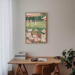 May include: A framed painting of three women wearing hats and sitting on chairs, watching a tennis match on a green court. The painting is in a light wood frame and is hanging on a white wall.
