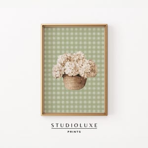 Op de afbeelding: Een ingelijste print met een aquarel illustratie van een mand vol witte hortensia's. De achtergrond is een lichtgroen ruitjespatroon. De lijst is goudkleurig en de tekst "STUDIOLUXE PRINTS" staat onderaan.