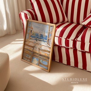 May include: A framed painting of a seascape with boats and a building in the distance. The painting is in a wooden frame and is sitting on a red and white striped couch.
