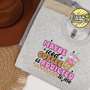 Puede incluir: Sudadera gris con el texto "Maybe iced coffee is addicted to me" en letras rosas y marrones. Un vaso de caf&eacute; helado de dibujos animados con una cara sonriente est&aacute; al lado del texto.