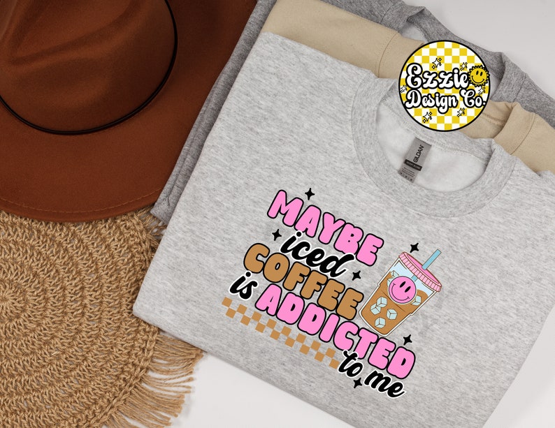 Puede incluir: Sudadera gris con el texto "Maybe iced coffee is addicted to me" en letras rosas y marrones. Un vaso de caf&eacute; helado de dibujos animados con una cara sonriente est&aacute; al lado del texto.